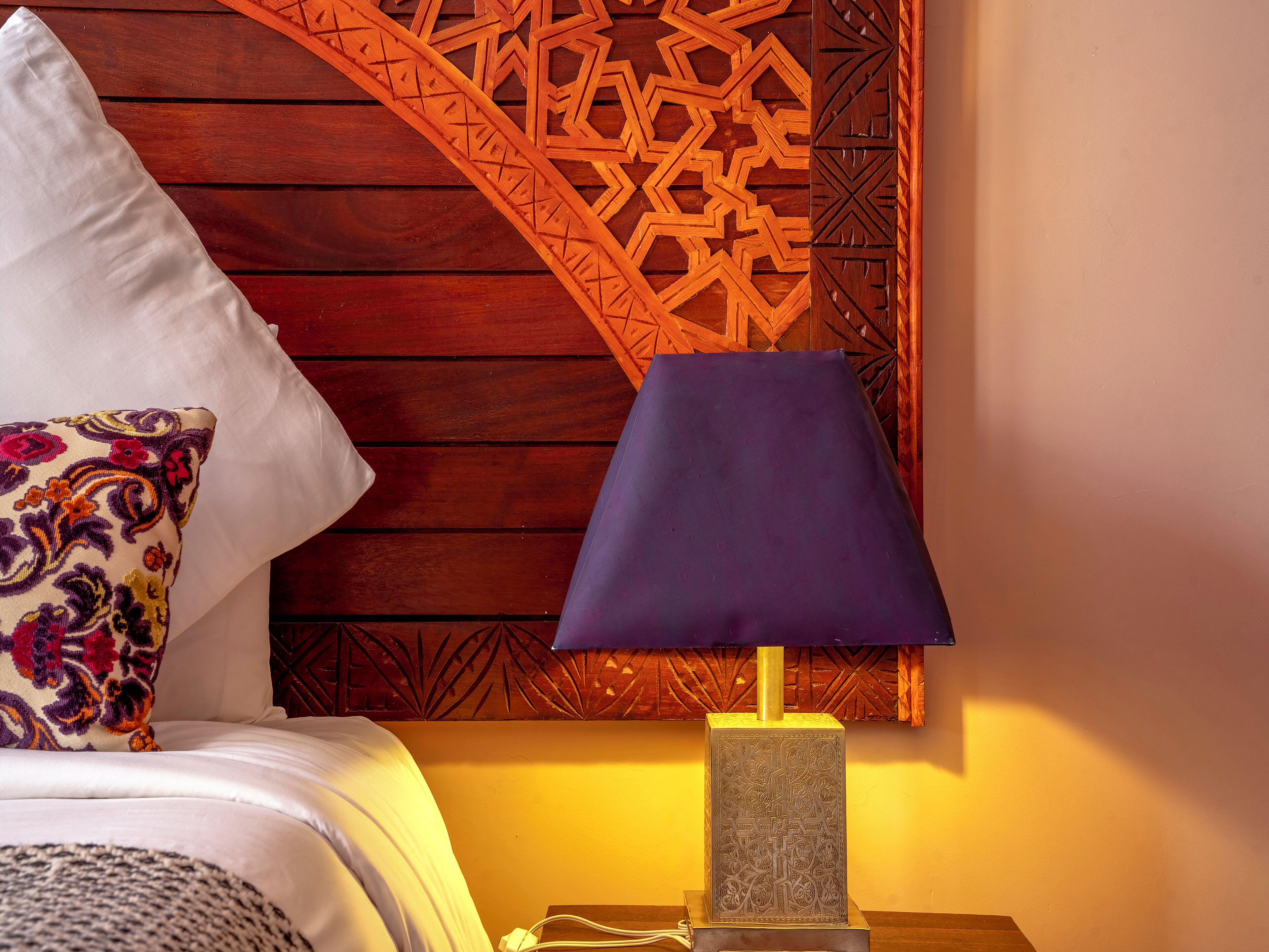 Una cama con ropa de cama blanca y una almohada colorida, una lámpara azul sobre una mesita de noche de madera, y una pared de madera con diseños geométricos.