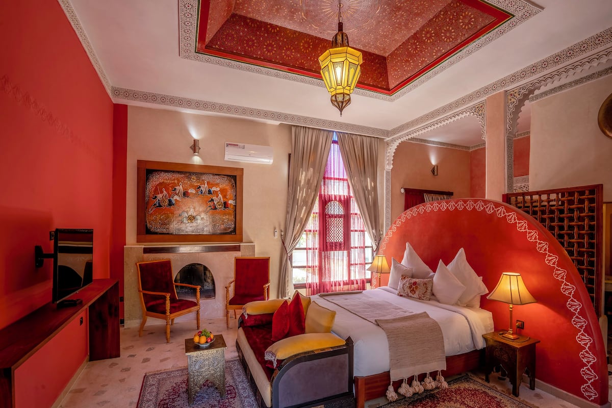A traditional Moroccan-style bedroom with a red and beige color scheme, a large bed with white pillows, a chandelier, and a fireplace.