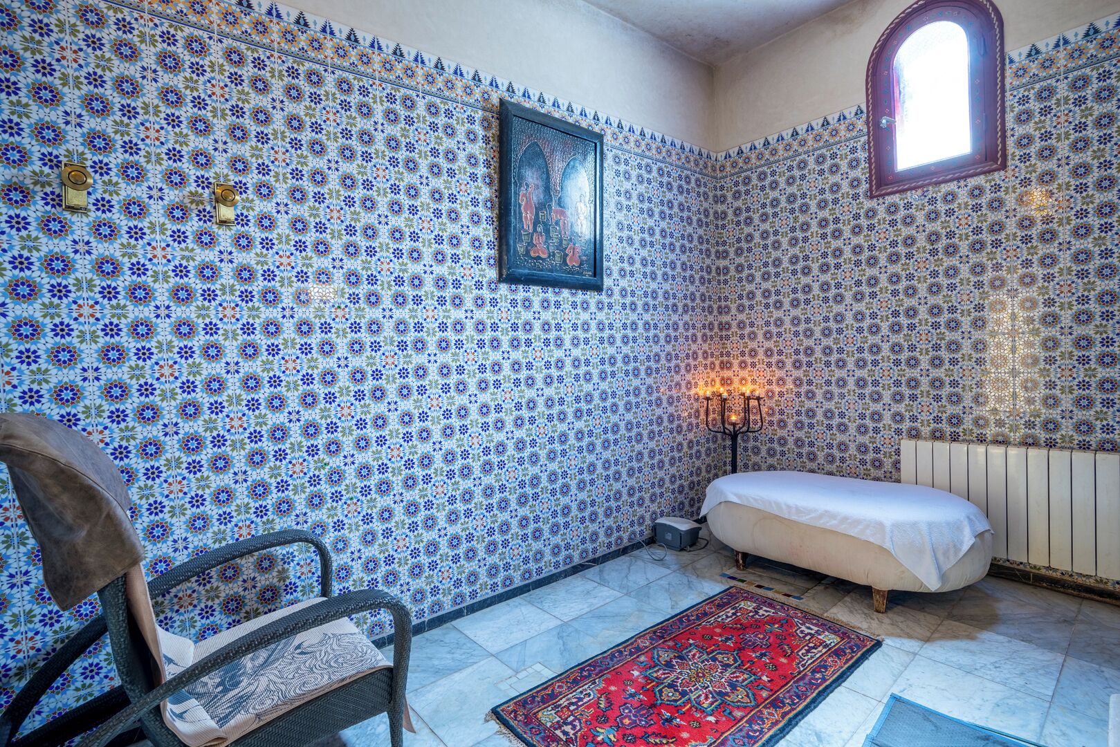 A room with patterned tile walls, a bed, a chair, a rug, and a candelabra.