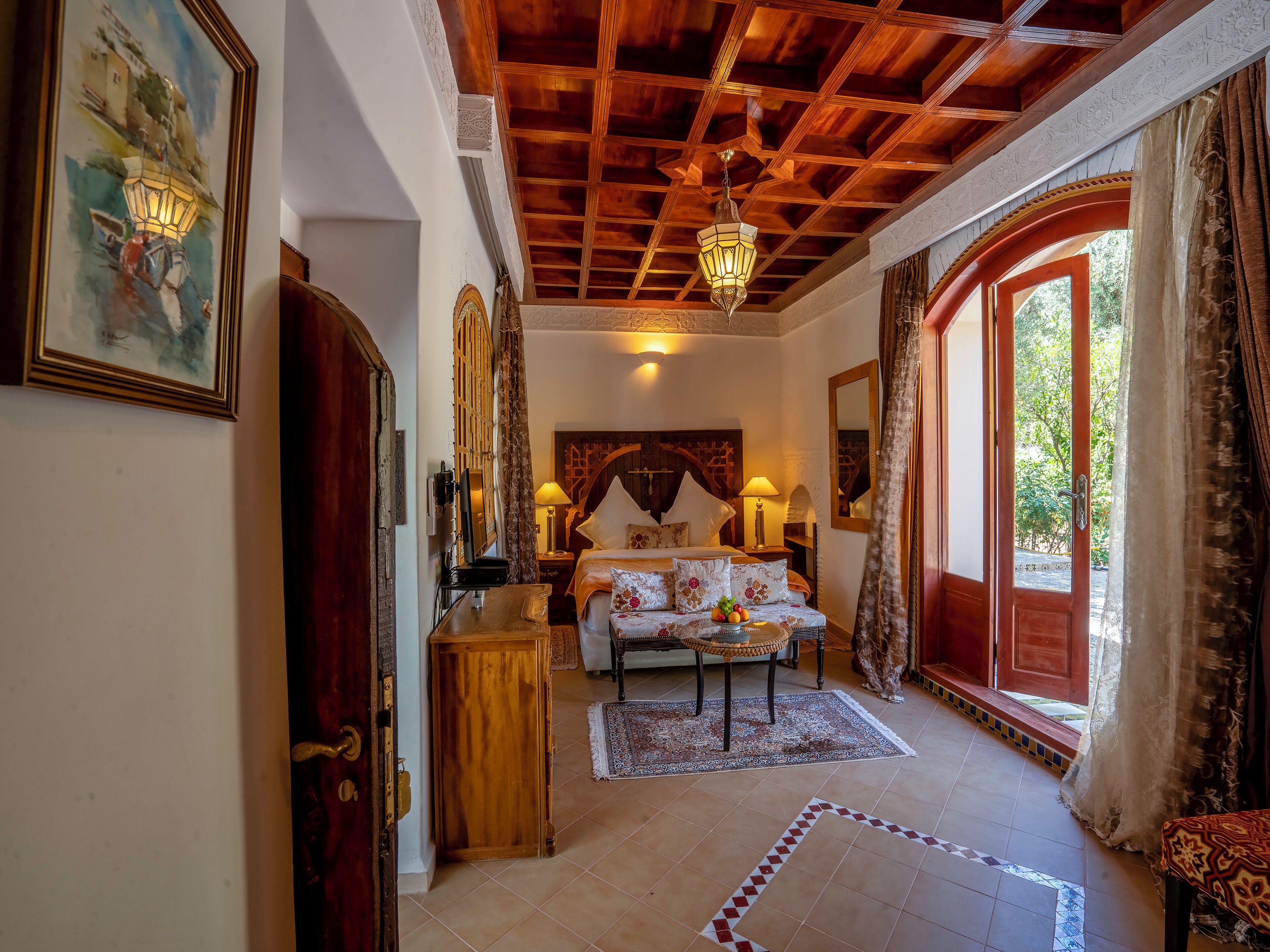 Habitación de hotel con techo de madera, cama con colcha estampada floral y escritorio con lámpara, con vistas a zonas verdes.