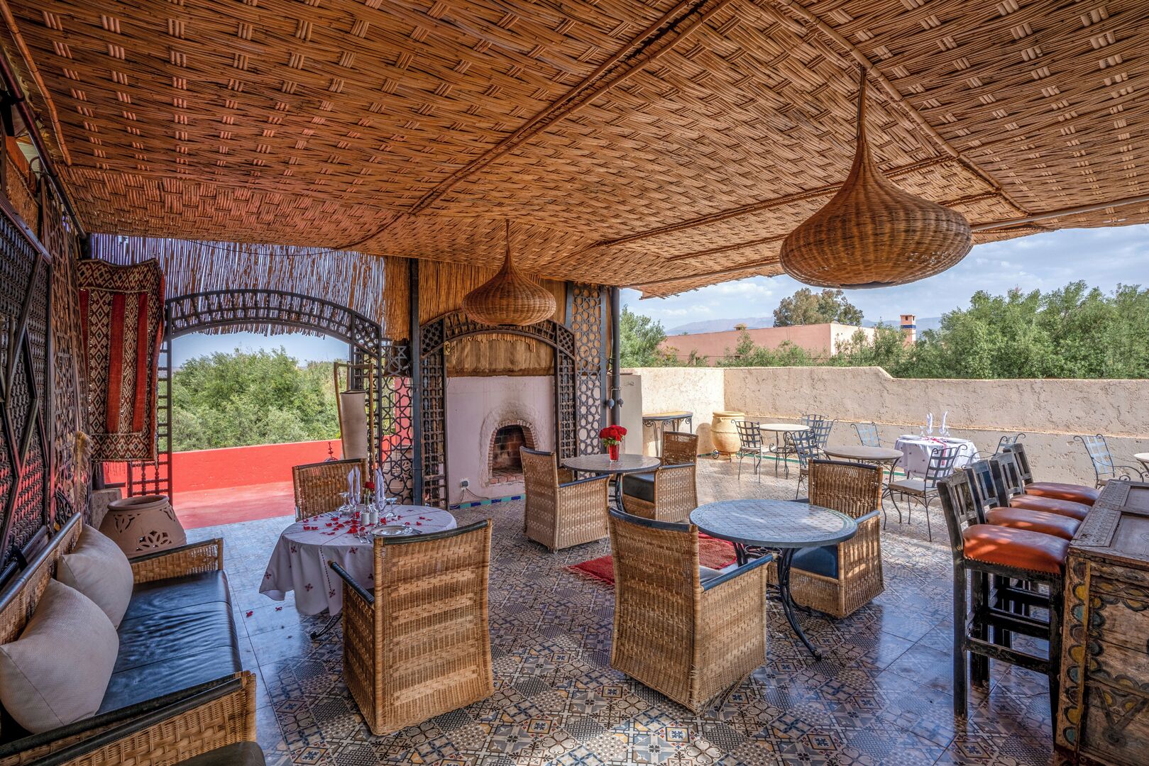 An outdoor patio with wicker furniture, a central table with a vase of flowers, and a view of greenery and buildings in the background.