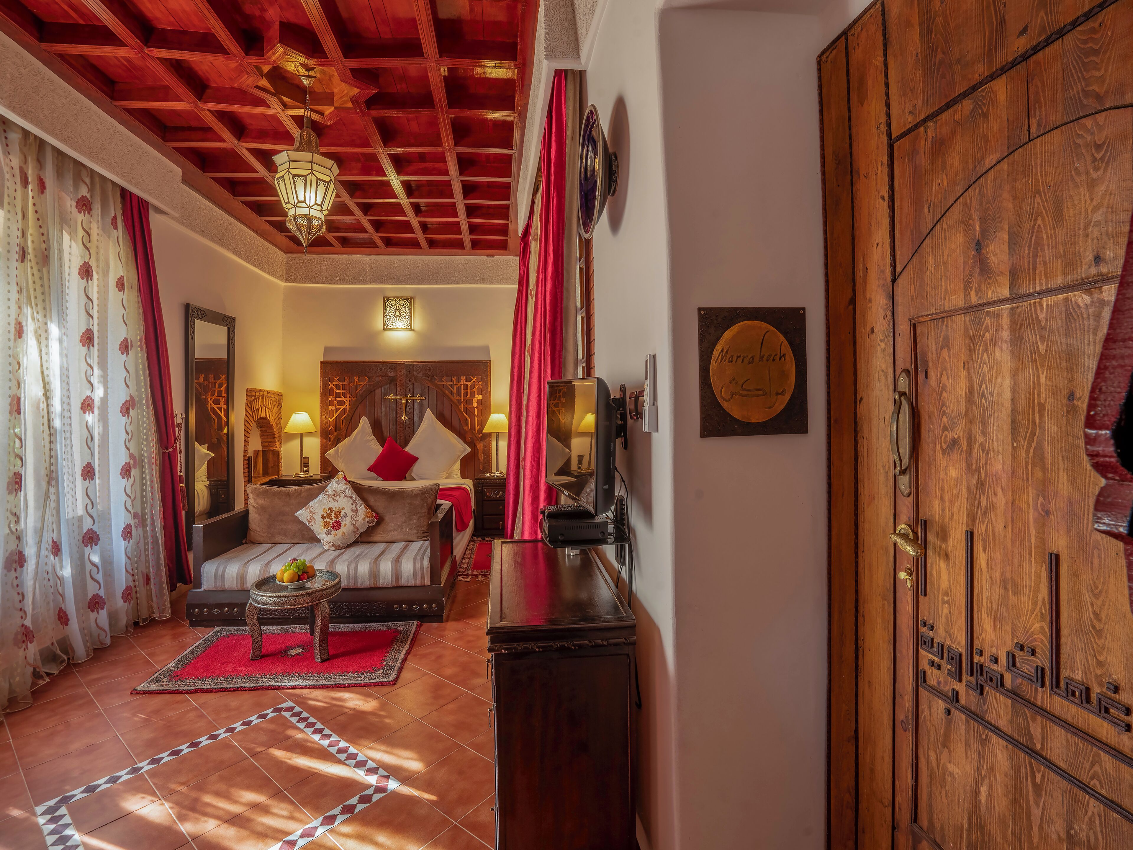 Habitació marroquí tradicional amb una porta de fusta, un llit amb coixins vermells i blancs, un armari de fusta i una catifes amb dibuixos vermells i blancs.
