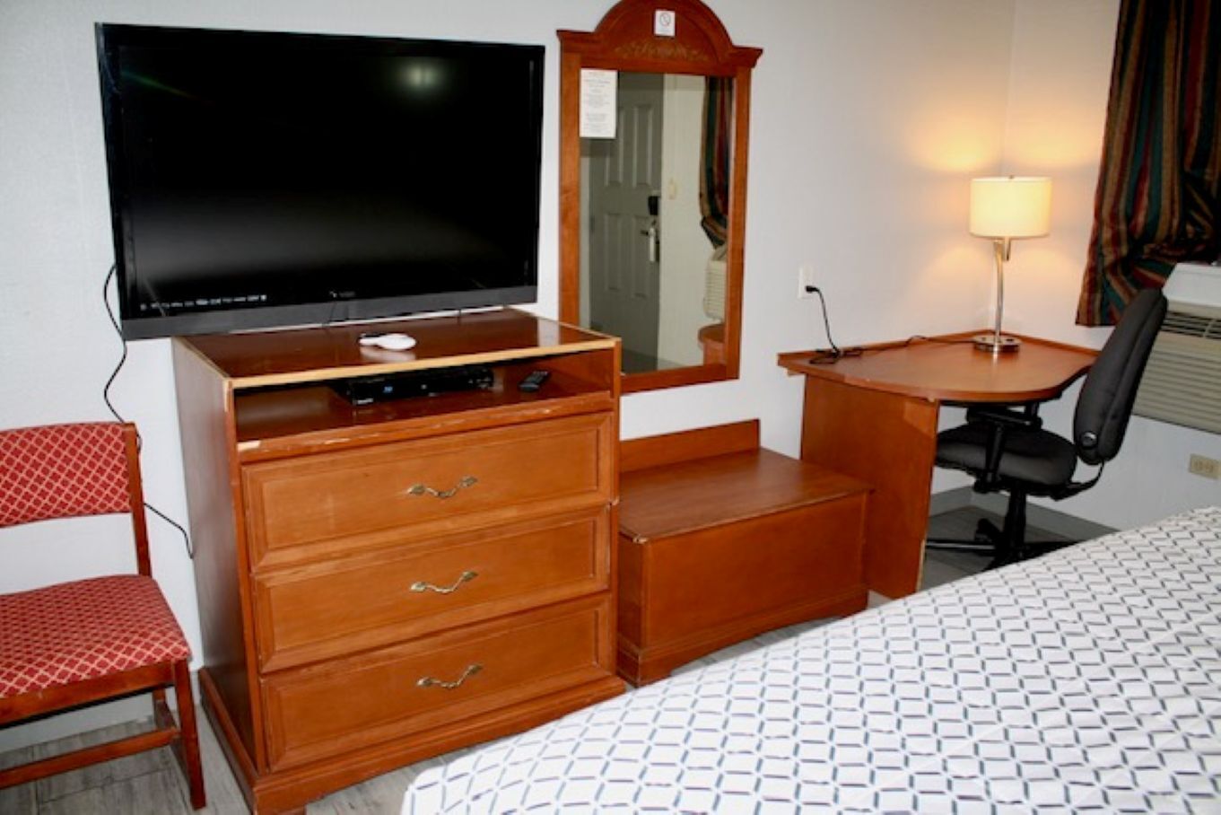 A hotel room with a bed, a wooden dresser with drawers, a desk with a chair, a flat-screen TV, a mirror, and a lamp.