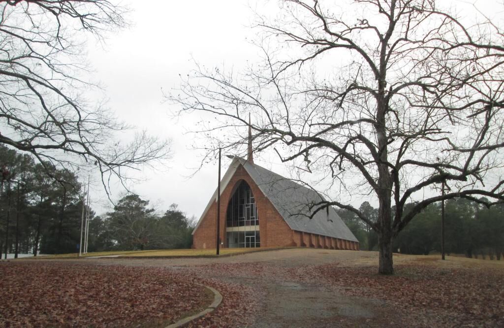A building with a triangular roof and large windows, surrounded by trees and a leaf-covered ground.