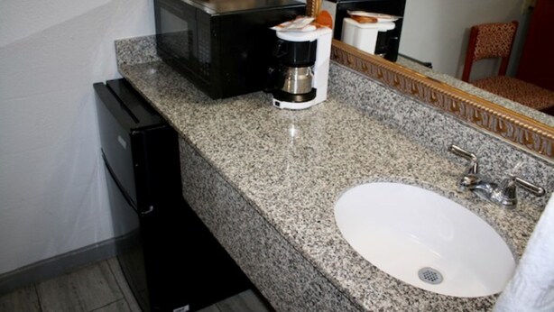 A bathroom with a granite countertop, a white sink, a black microwave, a coffee maker, and a mirror with a wooden frame.