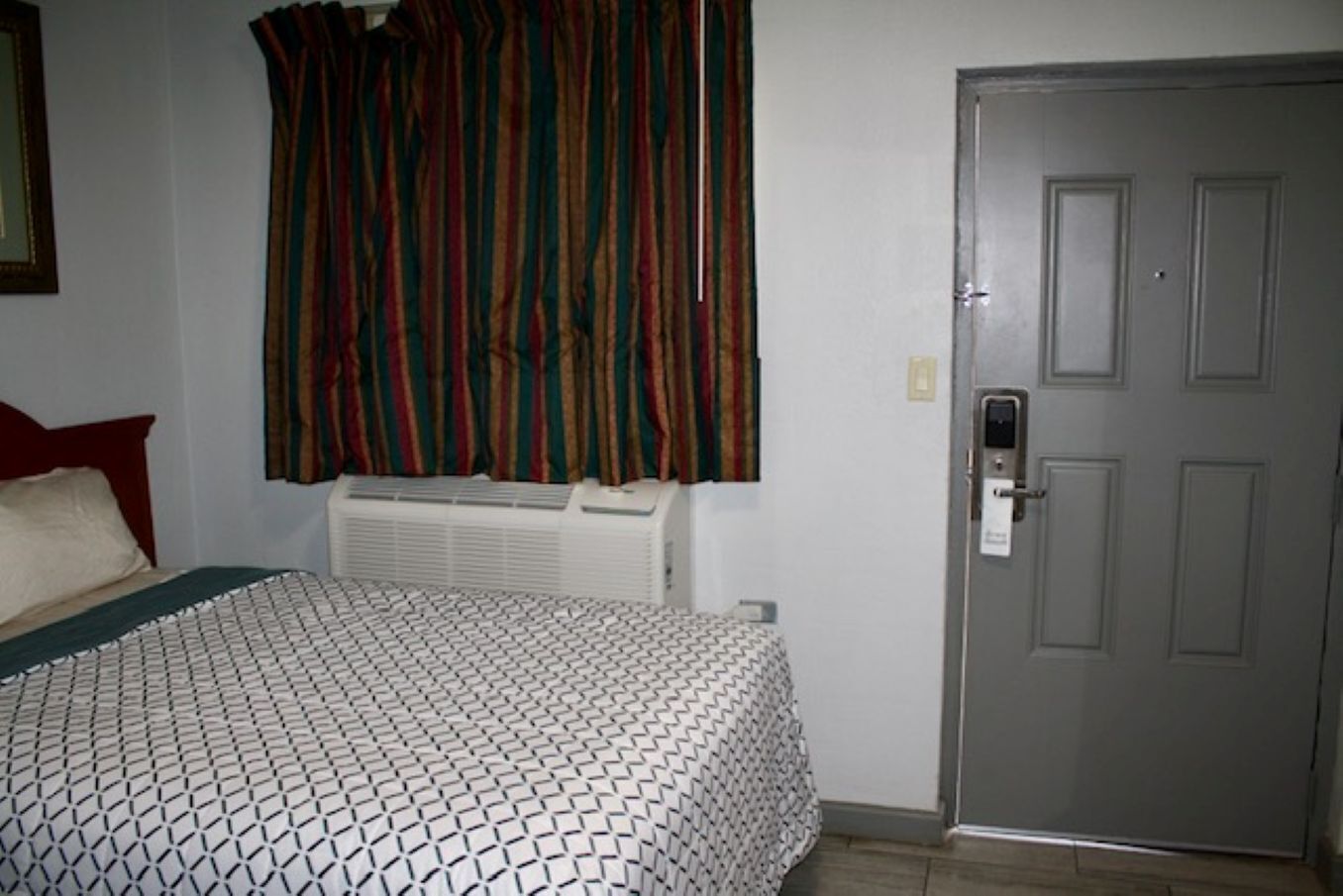 A hotel room with a bed, a window with curtains, a door, and an air conditioning unit.