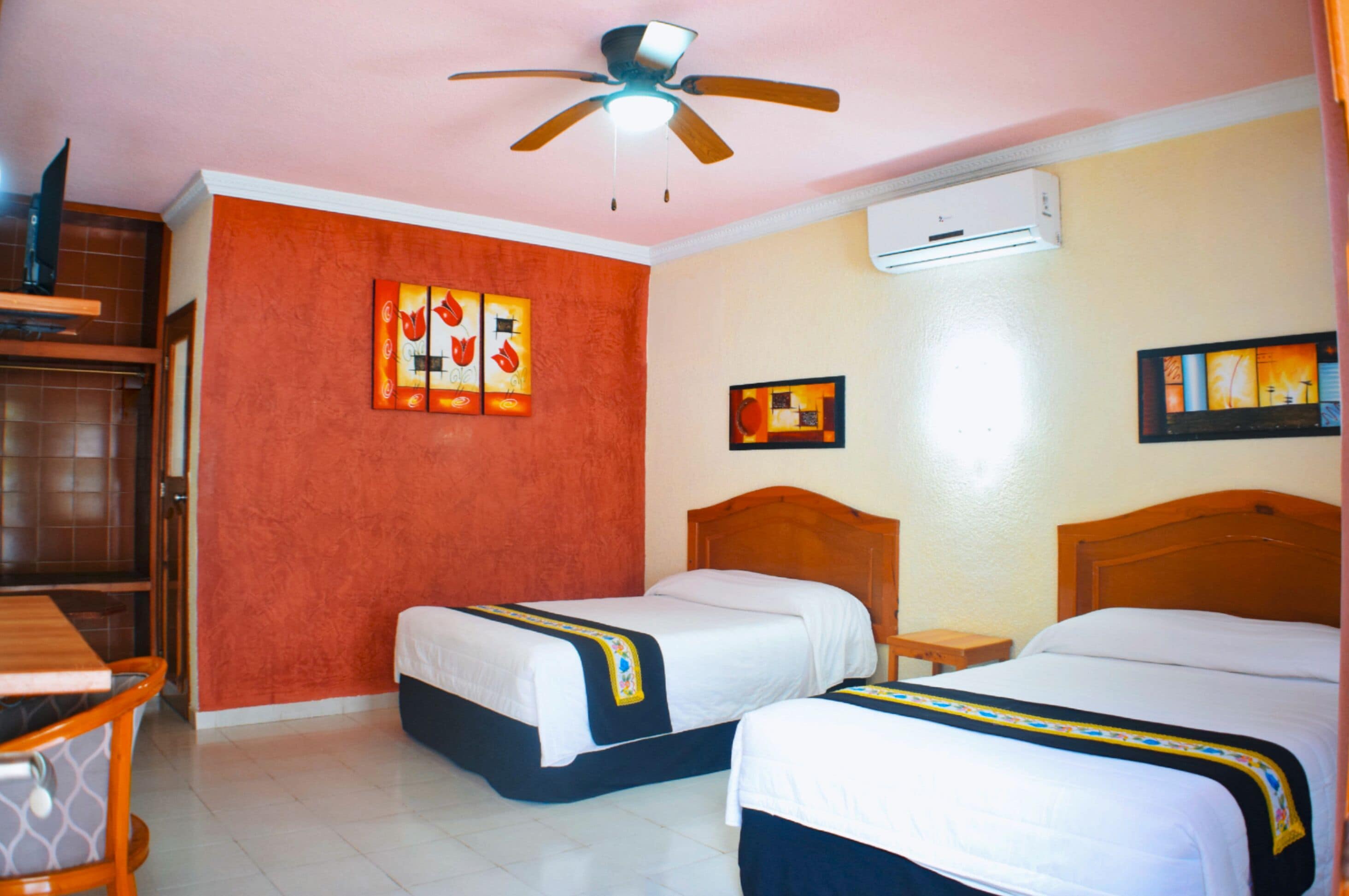 A hotel room with two beds, a ceiling fan, an air conditioning unit, and artwork on the walls.