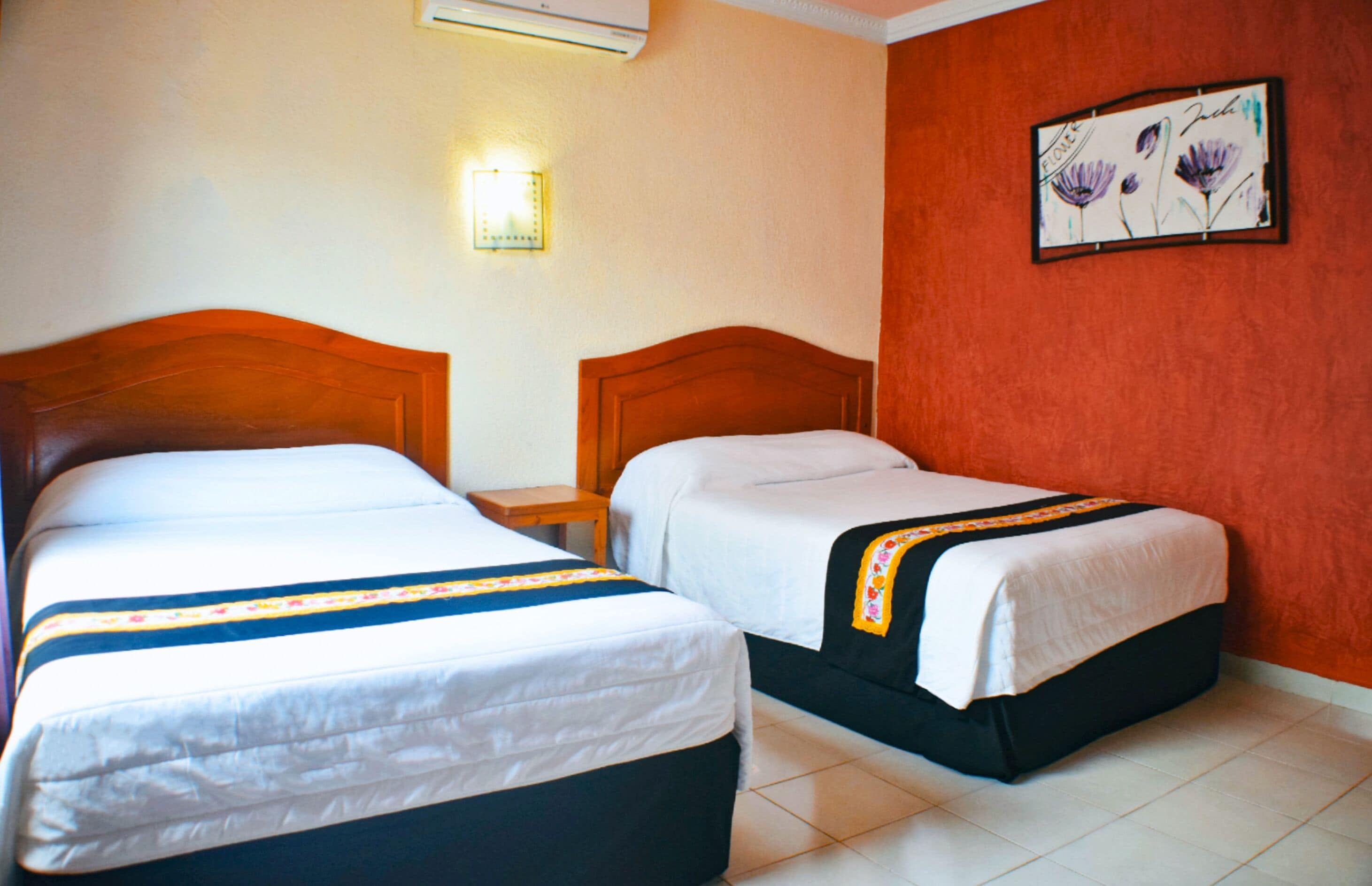A hotel room with two single beds, a nightstand, a wall-mounted lamp, and a floral artwork on the wall.