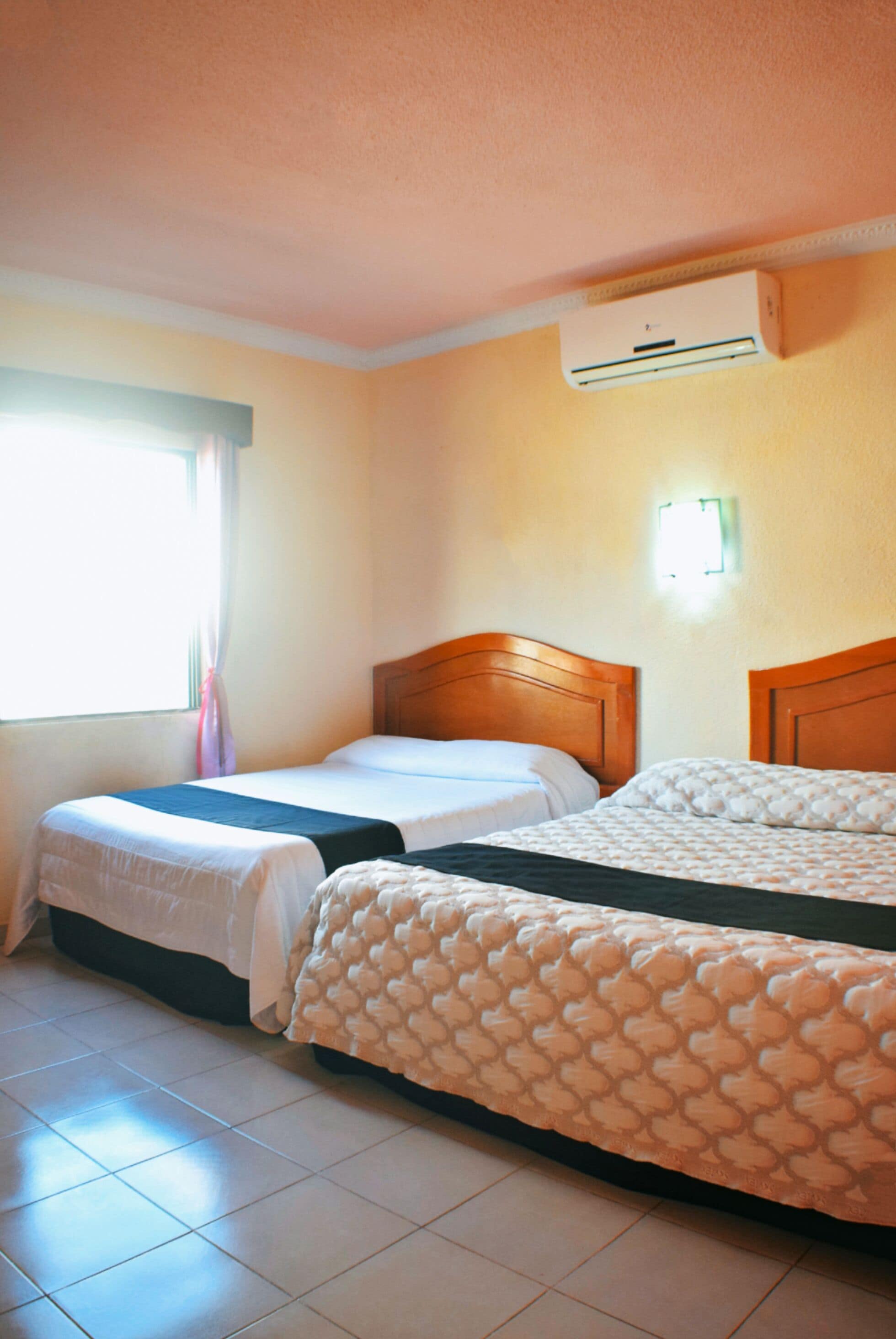 A hotel room with two single beds, a window with curtains, an air conditioning unit, and a wall-mounted light.