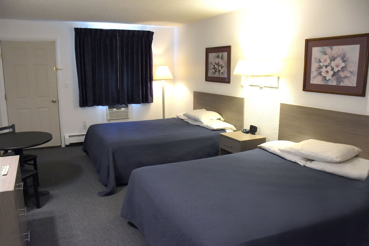 A hotel room with two beds, a desk, and a chair. There are two lamps on either side of the beds and a door on the left.