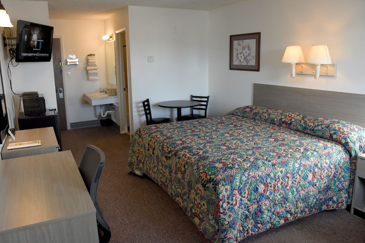 A hotel room with a bed, a desk, a chair, a small table, a TV, a sink, and two wall-mounted lamps.