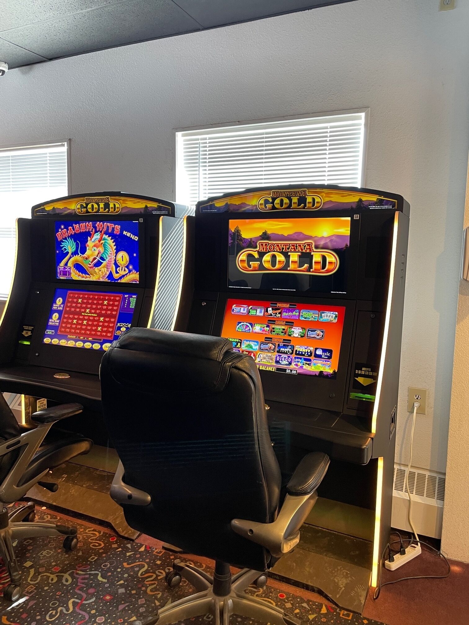 Two slot machines labeled "Montana Gold" with a variety of game screens, a black office chair, and a patterned carpet.