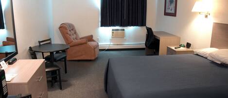 A hotel room with a bed, a desk, a chair, a desk lamp, a window with curtains, and a wall-mounted picture.