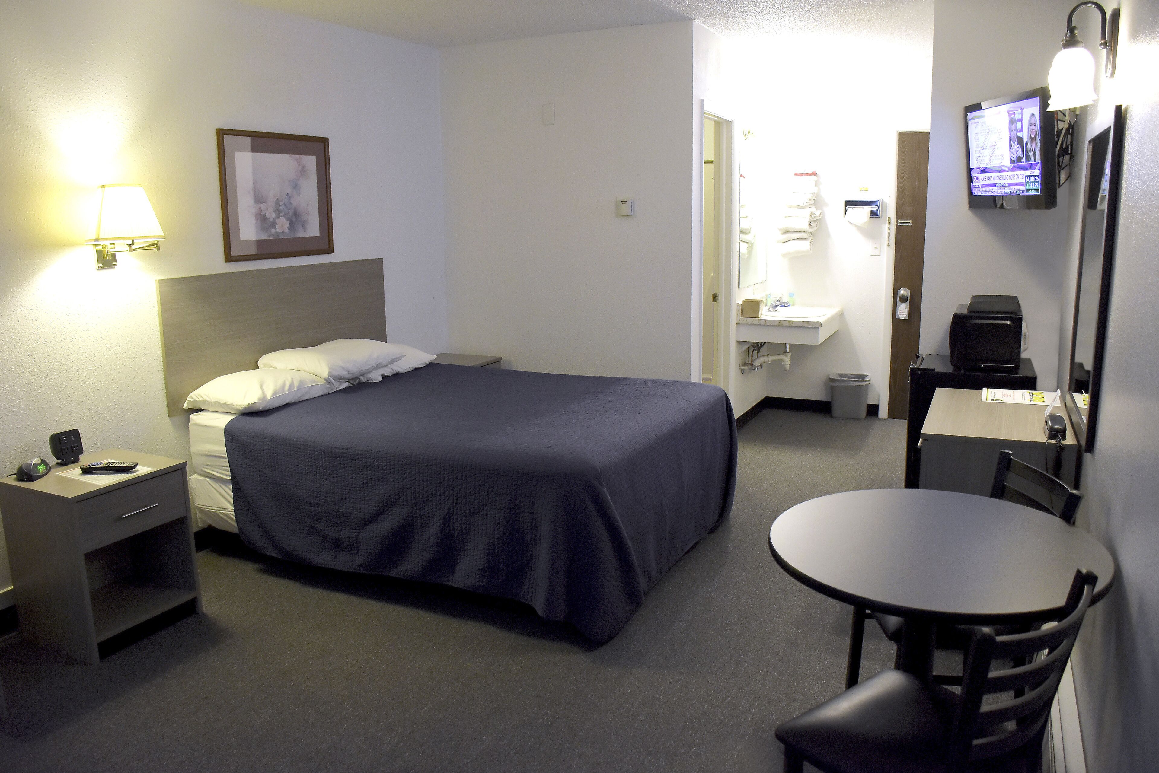 A hotel room with a bed, a desk with a chair, a small table, a TV, and a lamp.