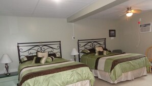 A hotel room with two beds, each with a metal frame and green bedding. There are bedside tables with lamps, a ceiling fan, and a wall-mounted air conditioning unit. - Happy Motel (Edmundston)