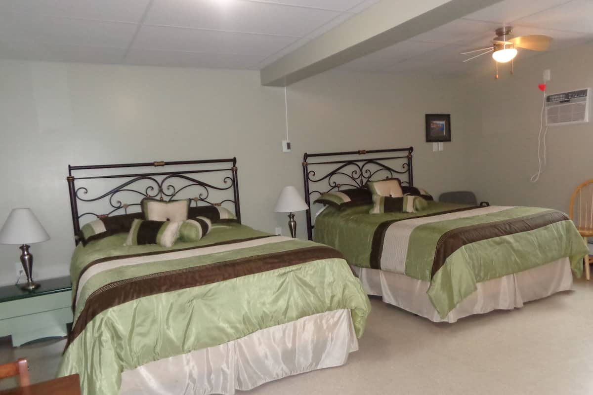 A hotel room with two beds, each with a metal frame and green bedding. There are bedside tables with lamps, a ceiling fan, and a wall-mounted air conditioning unit.
