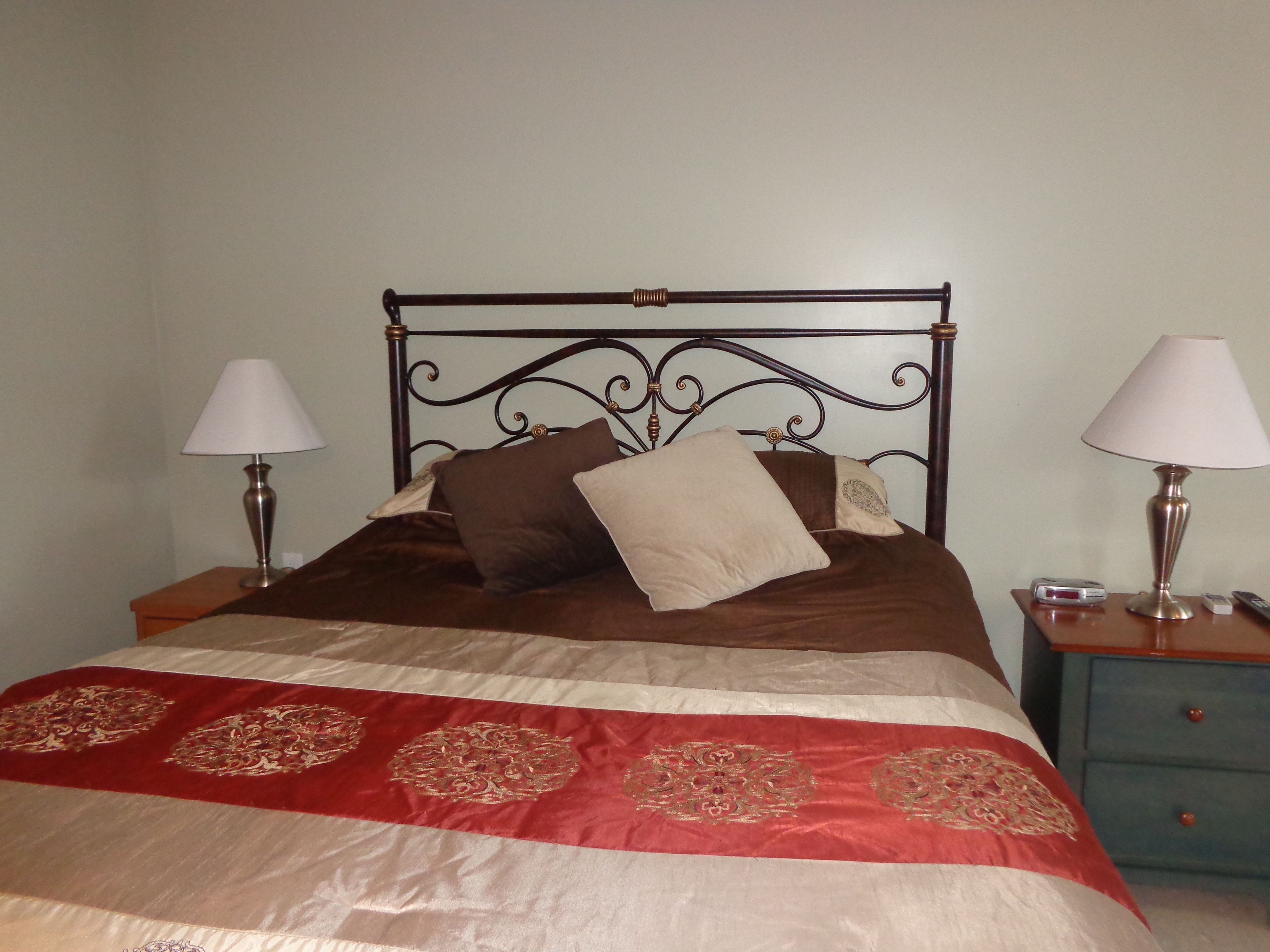 Un lit bien fait, recouvert d’une couvre-lit rouge et beige, avec deux lampes de chevet et une petite table de chevet sur laquelle se trouve une horloge.