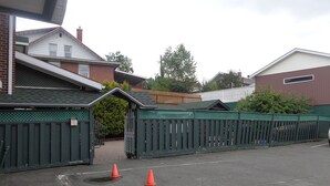 A gated parking area with two orange traffic cones, a green fence, and a brick building with a sloped roof. - Happy Motel (Edmundston)