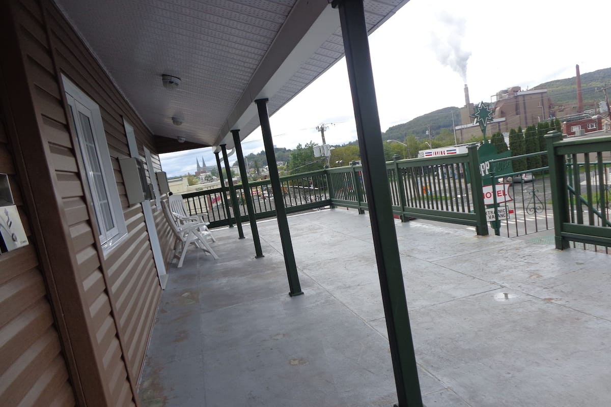 A covered outdoor area with a concrete floor, metal railings, and a view of a hotel and industrial buildings in the background.