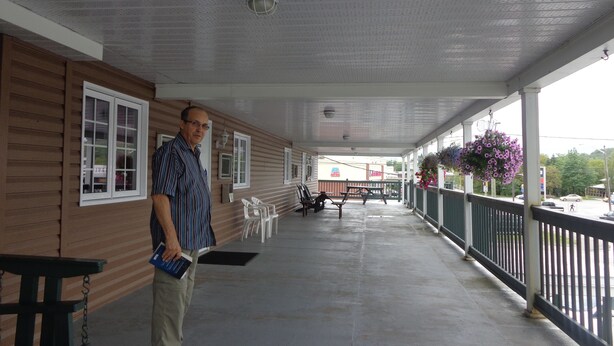 A covered porch with a man standing, wooden walls, white-framed windows, outdoor seating, and hanging flower baskets. - Happy Motel (Edmundston)