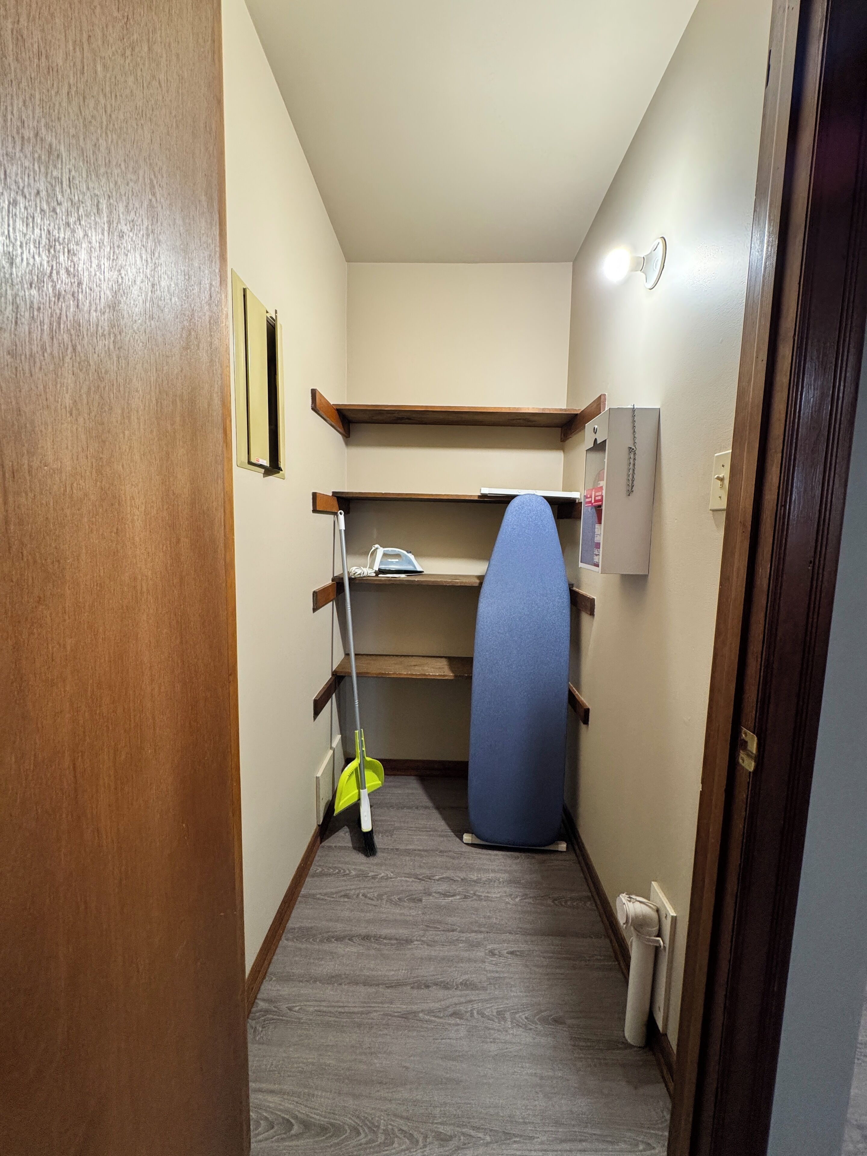 Un couloir étroit avec une porte en bois sur la gauche, une planche à repasser bleue, un balai-serpillière et une étagère avec un miroir et un téléphone.