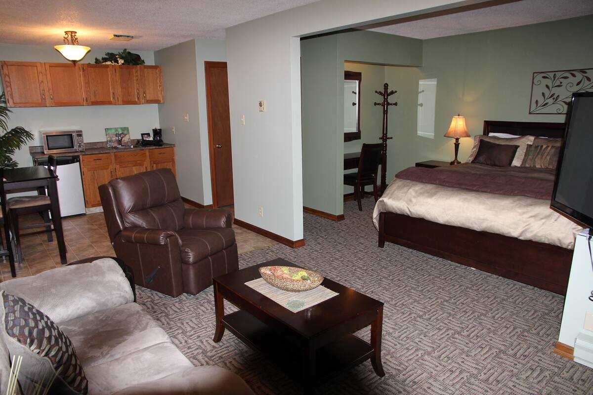 Uma sala de estar com poltrona de couro marrom, sofá, mesa de centro de madeira e cama com cabeceira marrom.