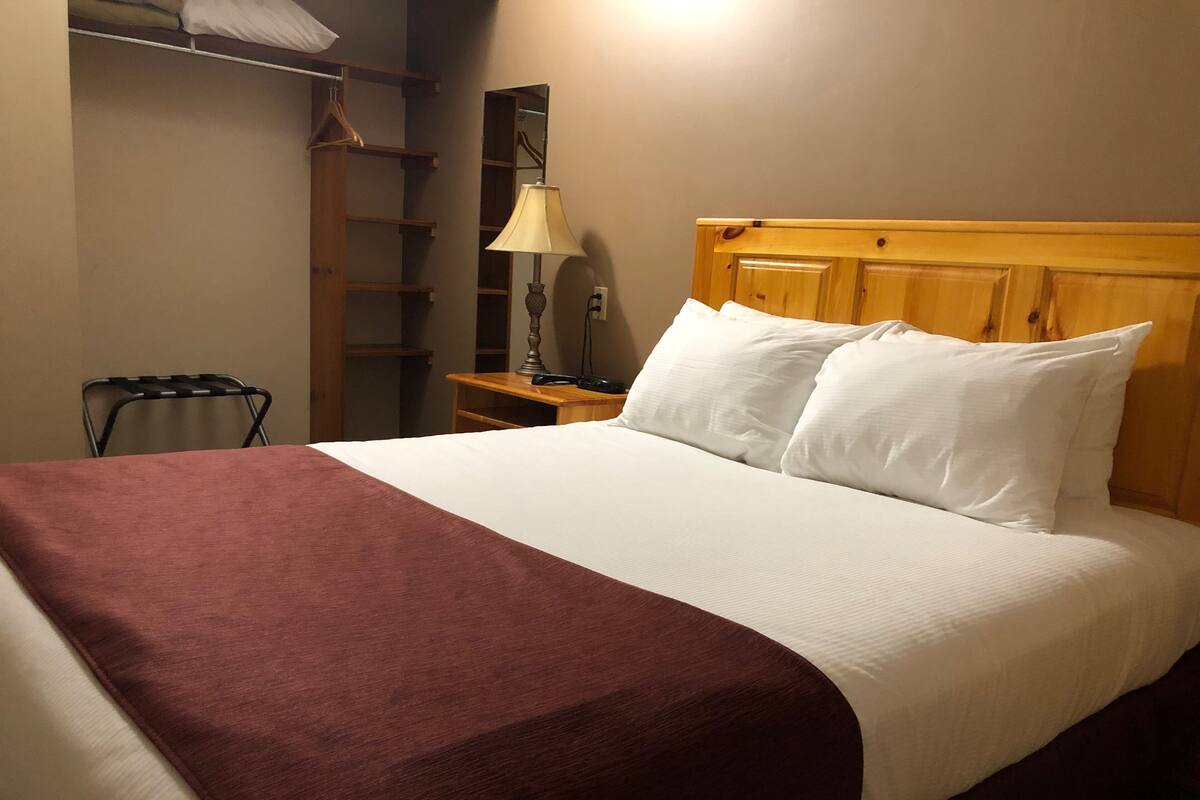 Cama bem arrumada com cabeceira de madeira, mesa de cabeceira com abajur e guarda-roupa com cabides.