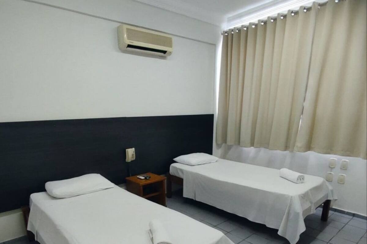 A hotel room with two single beds, a bedside table with a phone, an air conditioning unit, and a window with curtains.