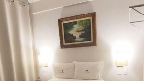 A neatly made bed with a rolled towel, a painting above it, and a small stool beside the bed. - Titao Plaza Hotel (Campina Grande)