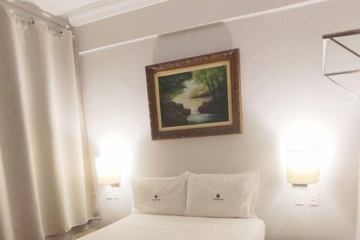 A neatly made bed with a rolled towel, a painting above it, and a small stool beside the bed.