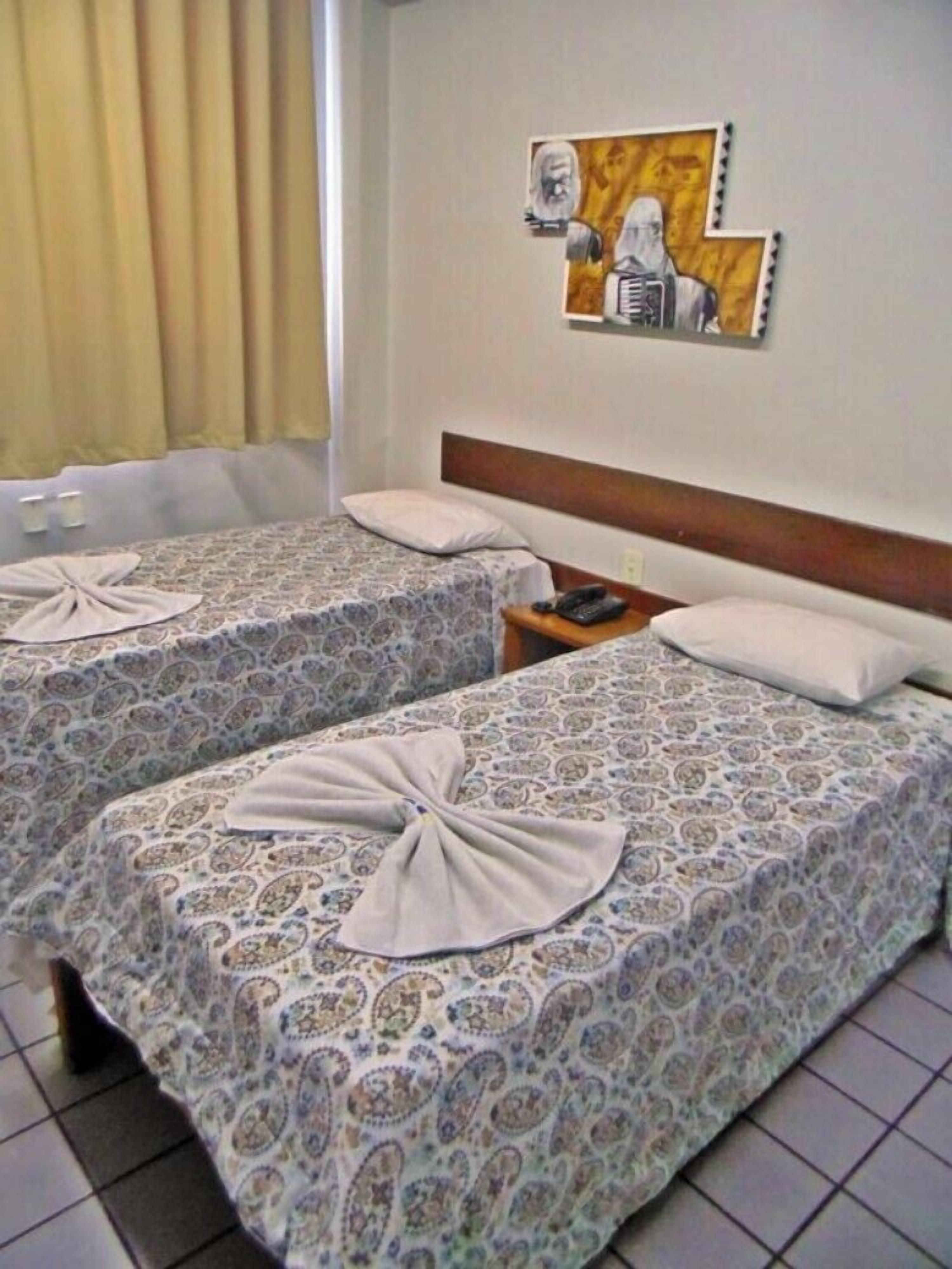 Habitación de hotel con dos camas, cada una con una colcha estampada y una toalla doblada en lazo. Se observa un mesita de noche con un teléfono y una obra de arte colgada en la pared sobre las camas.