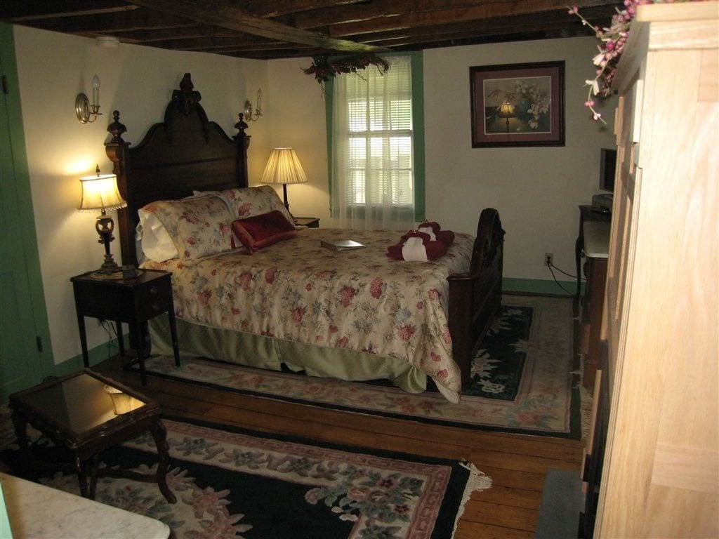 A bedroom with a wooden bed, floral bedding, a nightstand with a lamp, a small table, and a window with a green frame.