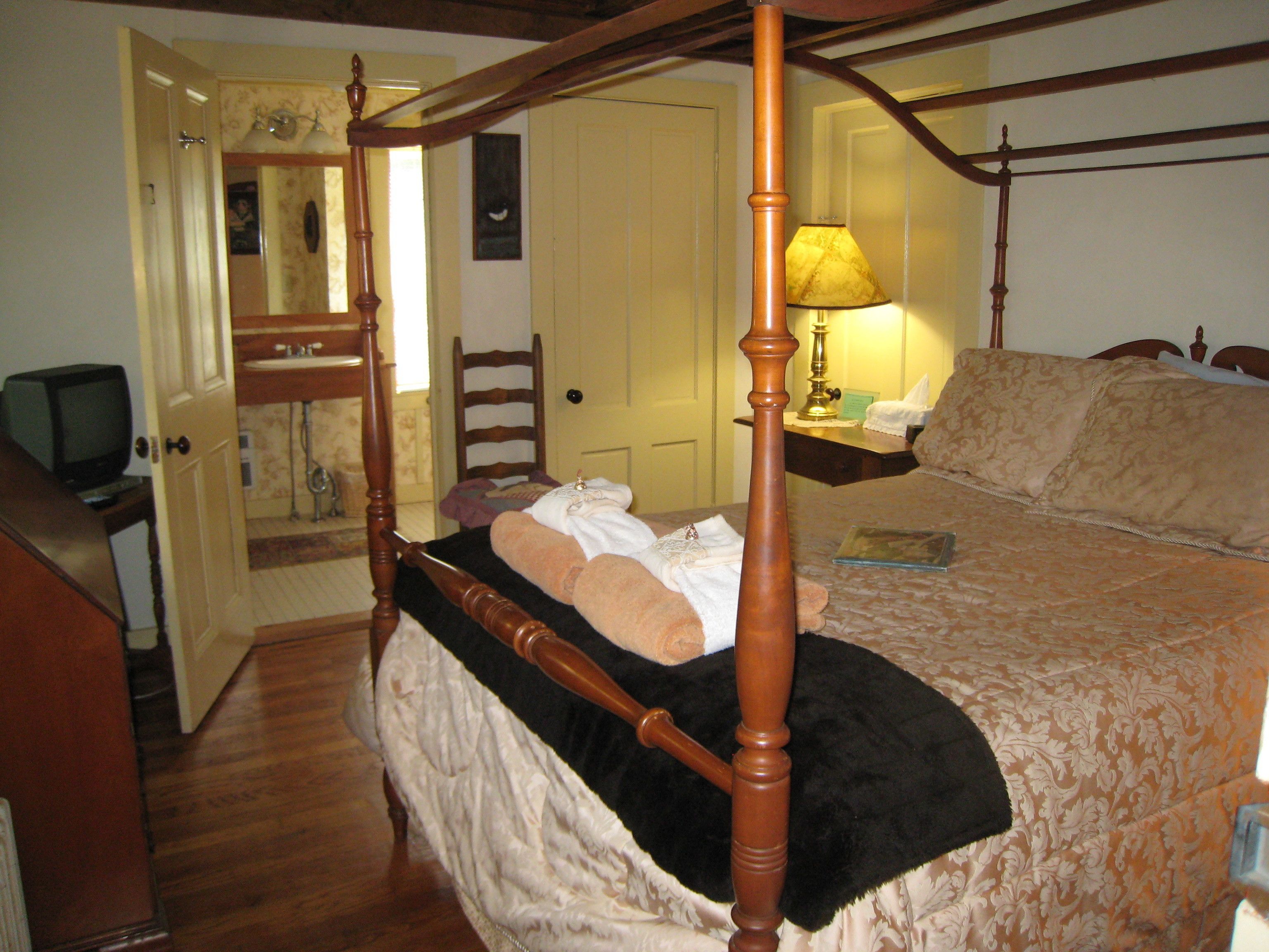A bedroom with a four-poster bed, a bedside table with a lamp, a chair, and a television in the corner.