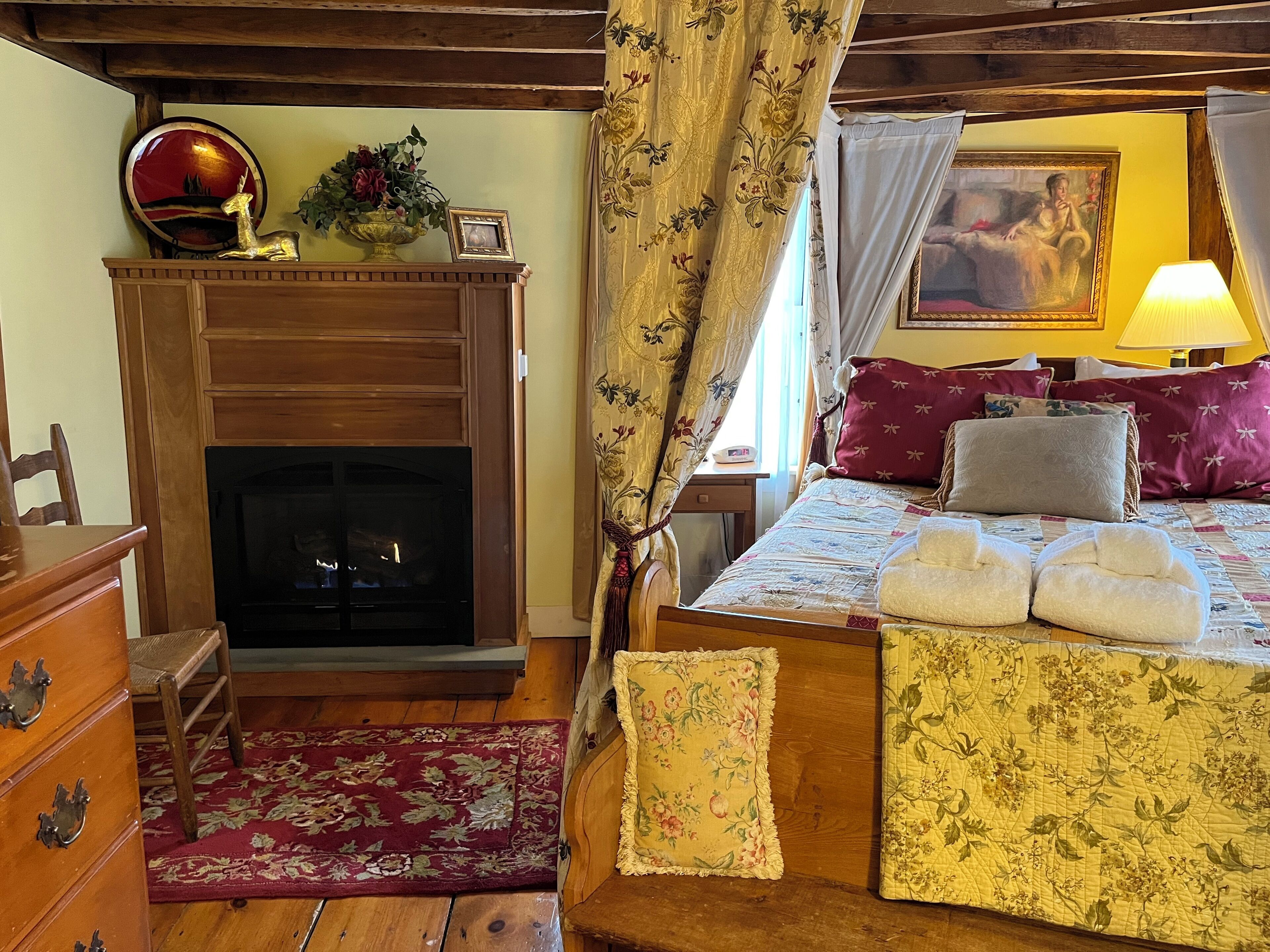 A cozy room with a fireplace, a bed with floral bedding, a wooden dresser, and a red rug.