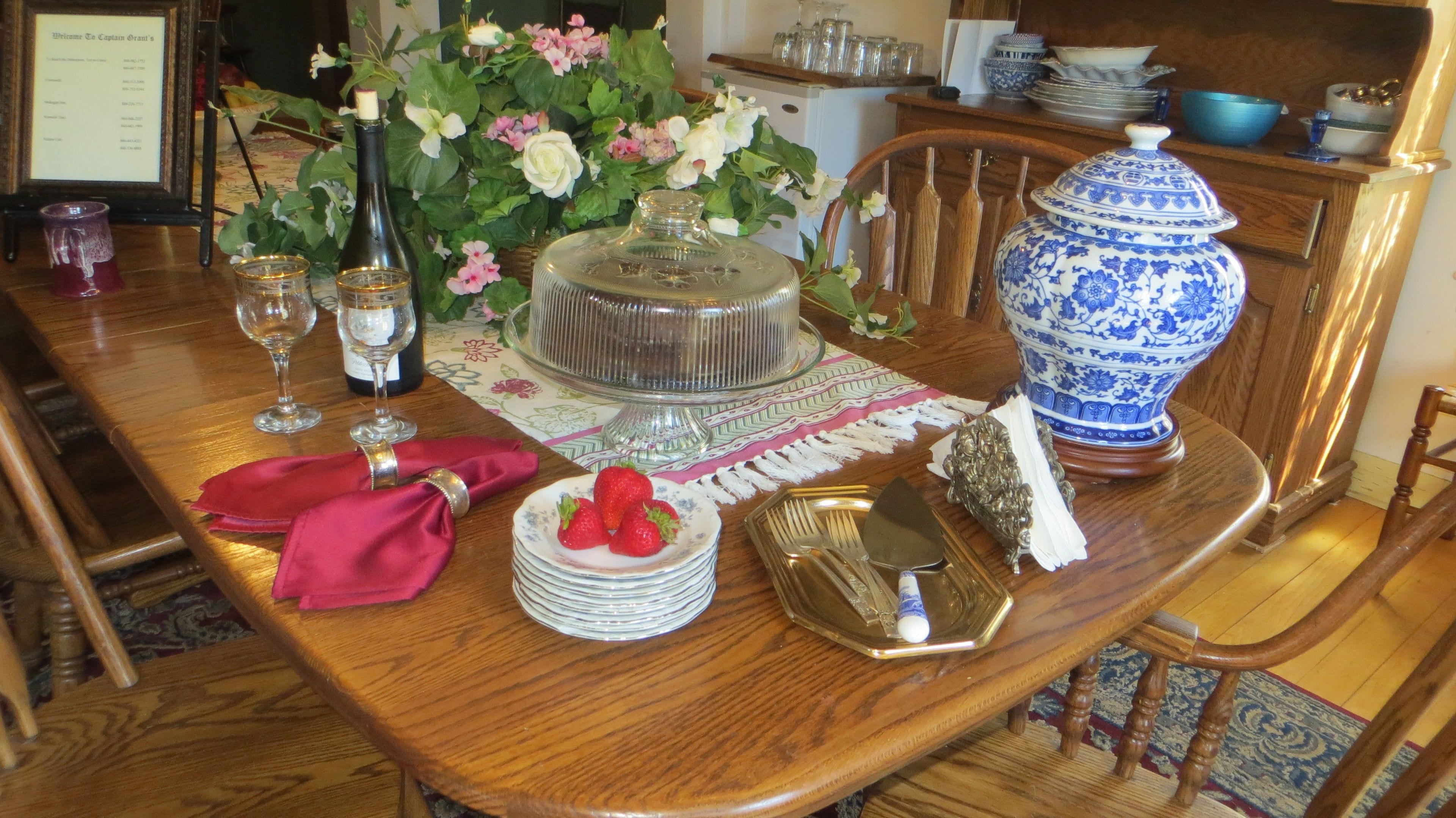 Un tavolo da pranzo in legno apparecchiato con piatti, posate, una torta, un vaso con fiori e una brocca di ceramica bianca e blu.