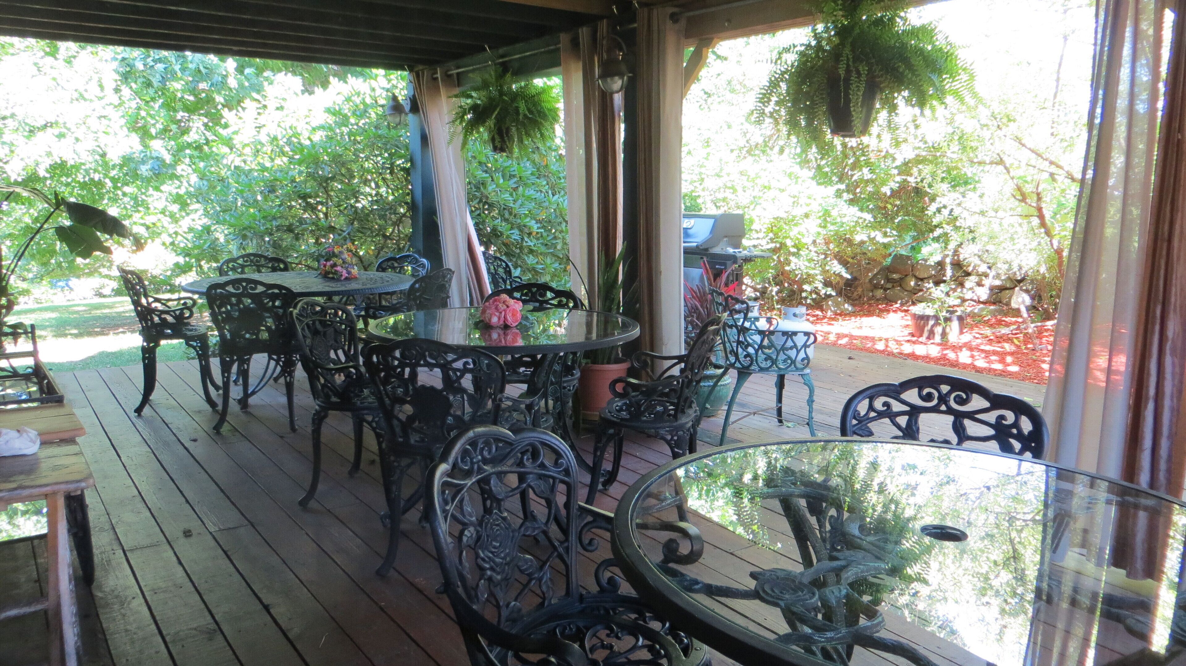Un patio al aire libre con una mesa redonda de vidrio rodeada de sillas de metal ornamentadas, una mesita redonda con un centro de mesa floral y plantas colgantes.
