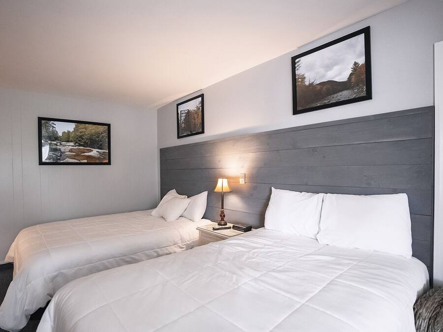 A hotel room with two beds, a wooden headboard, a nightstand with a lamp, and two framed pictures on the wall.