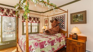 A bedroom with a four-poster bed, floral window valances, a large stuffed moose, a quilted bedspread, a nightstand with a lamp, and a framed landscape painting. - The Inn at Buck Hollow Farm (Fairfax)