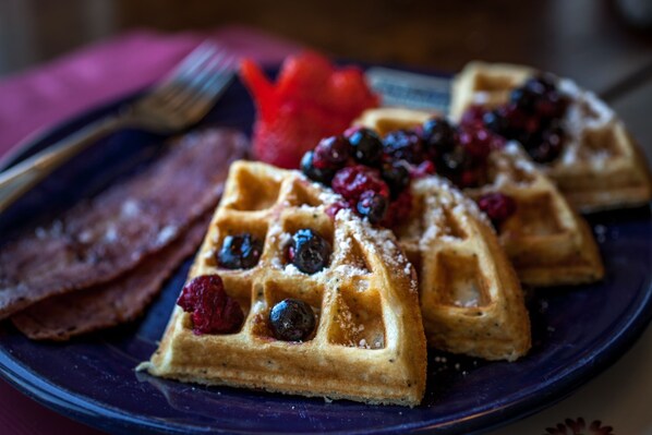 Waffles topped with berries and powdered sugar, served on a plate with a fork and a piece of bacon.