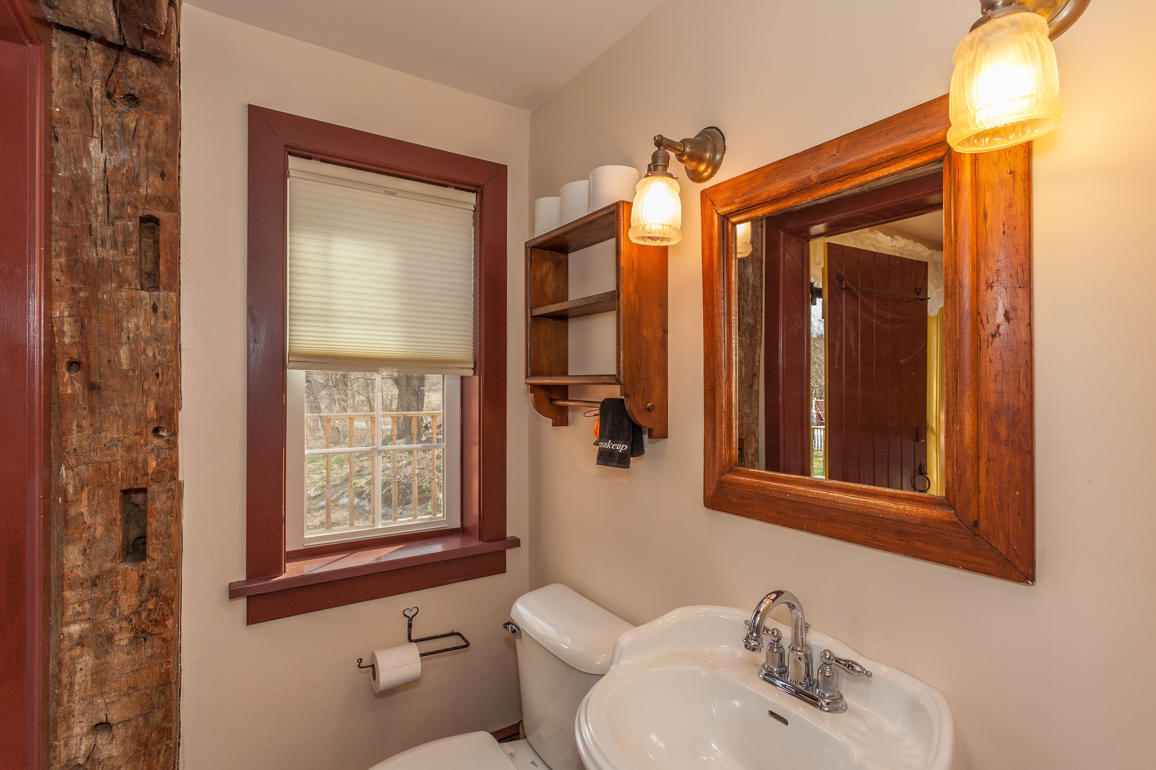 Un baño con inodoro, lavamanos, estantes de madera con artículos de aseo, una ventana con persianas y un espejo con luz.