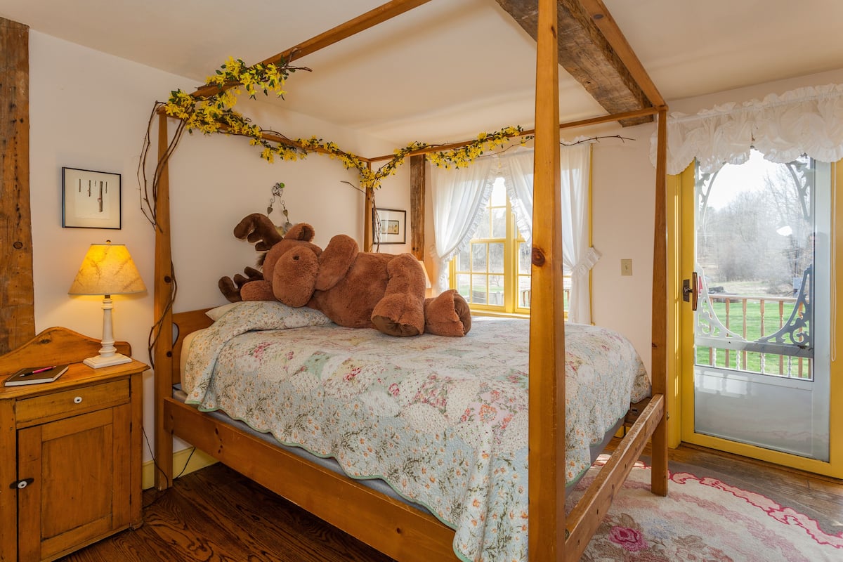 Ein Schlafzimmer mit einem Himmelbett, floralem Bettzeug, einem Nachttisch mit Lampe und Blick auf einen Garten durch eine Glastür.