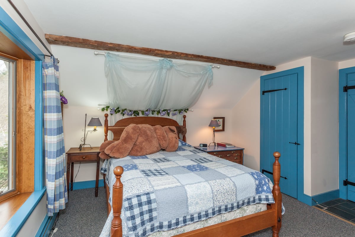 Ein Schlafzimmer mit einem hölzernen Bett, einem Fenster mit Vorhängen, einem Nachttisch mit Lampe und einer blauen Tür.