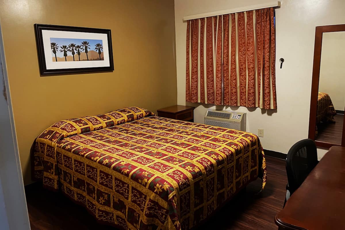 A bedroom with a bed, an air conditioner, a desk, a chair, a framed picture on the wall, and a window with curtains.