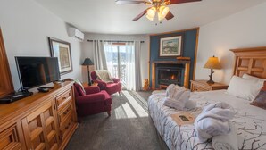 A hotel room with a bed, a TV, a fireplace, a ceiling fan, and a window with curtains. - Heather Lodge (Minden Hills)