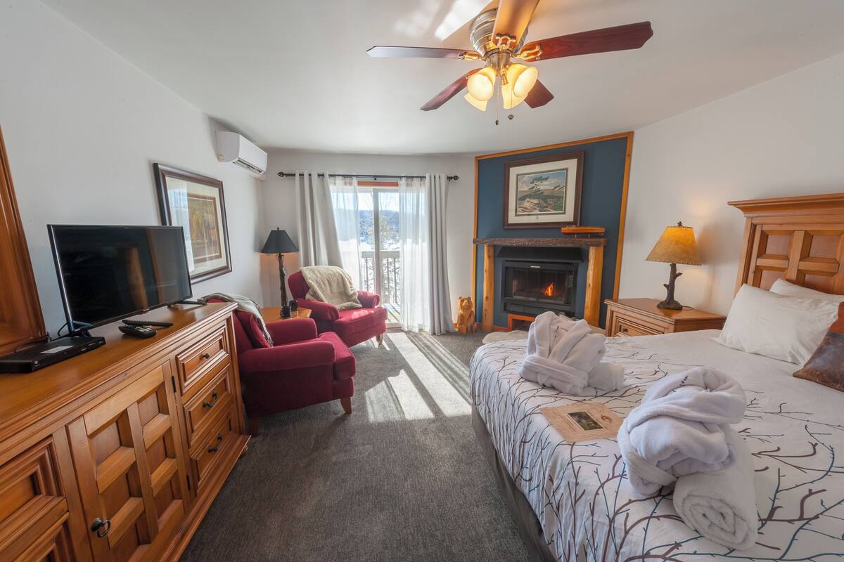 A hotel room with a bed, a TV, a fireplace, a ceiling fan, and a window with curtains.