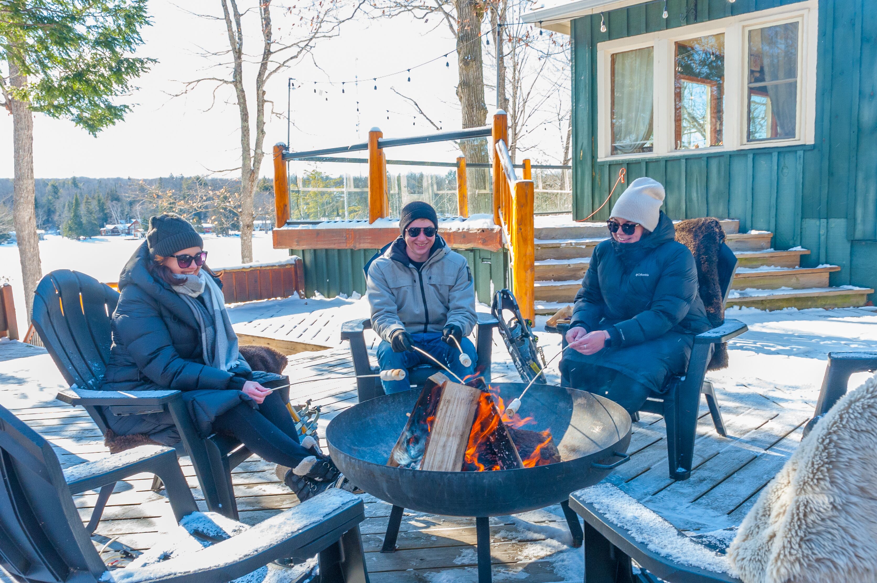 Three people sitting around a fire pit on a wooden deck with snow on the ground, a building with large windows in the background, and a wooden structure with a railing.