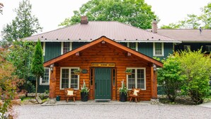 A wooden lodge with a green door, labeled "Office - Dining Room," featuring two wooden chairs and potted plants in front. - Heather Lodge (Minden Hills)