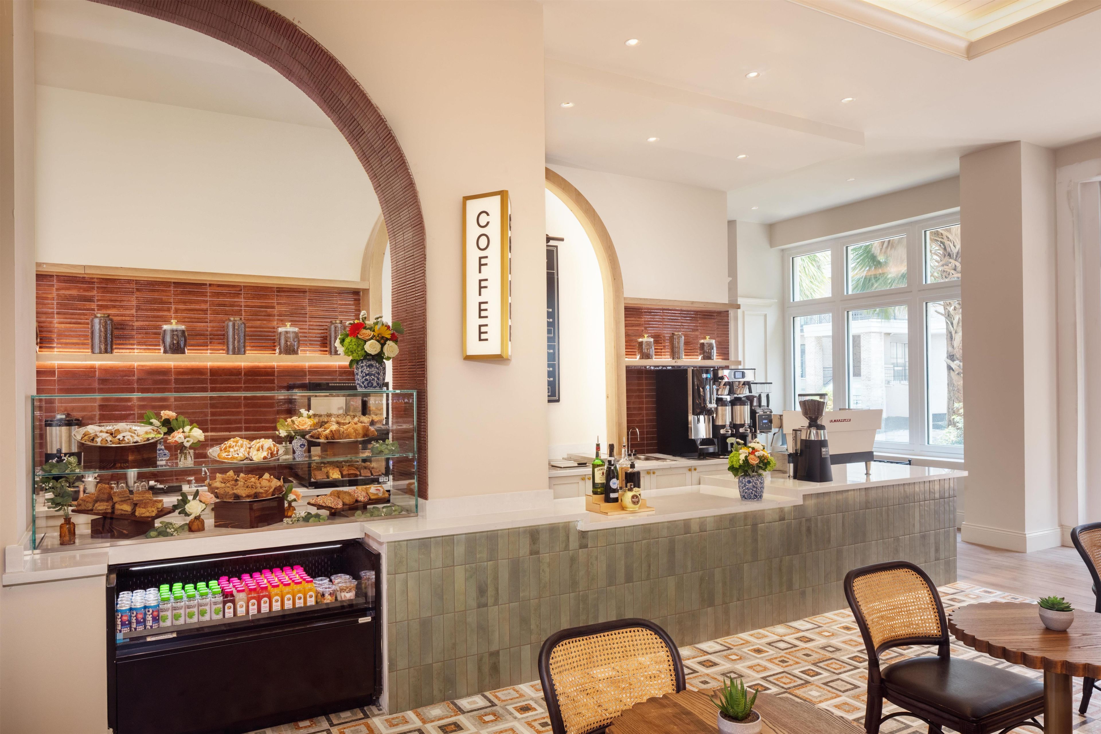 Um café com placa, vitrine refrigerada com bebidas e área de estar com cadeiras e mesa.