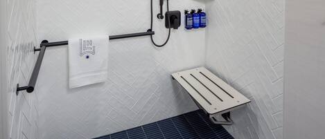 A bathroom with a white tiled wall, a blue tiled floor, a towel rack with a towel, a showerhead, and a shelf with bottles.