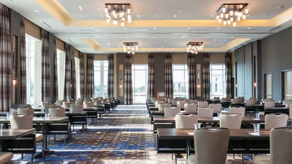 Un amplio salón de conferencias con filas de mesas y sillas, ventanas grandes y candelabros modernos.